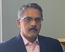 Person with short grey hair and a dark moustache wearing glasses, a pink button up shirt and a dark jacket.