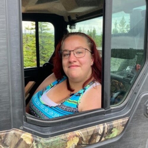 A woman with red hair and glasses is smiling while sitting in the passenger seat of a vehicle. She's wearing a colorful, patterned tank top, and the car door features a camouflage design. The background shows a scenic view with trees and a partly cloudy sky.