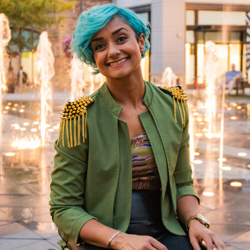 A person with short blue hair, wearing a green jacket with gold shoulder decorations and black pants, smiles while sitting in a wheelchair. They are positioned in front of an urban water fountain, with tall buildings and trees illuminated in the background.