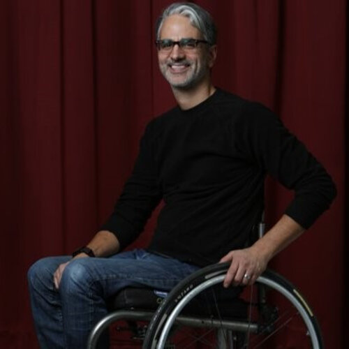 A smiling man with gray hair and glasses sits in a wheelchair. He's wearing a black long-sleeve shirt and blue jeans. The background consists of deep red curtains.