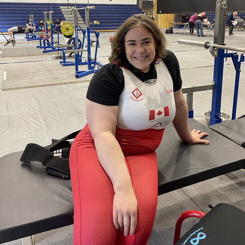 A smiling woman in a white shirt with a red maple leaf design and red pants sits on a bench in a gymnasium. Weightlifting equipment is seen in the background. She appears relaxed and confident.