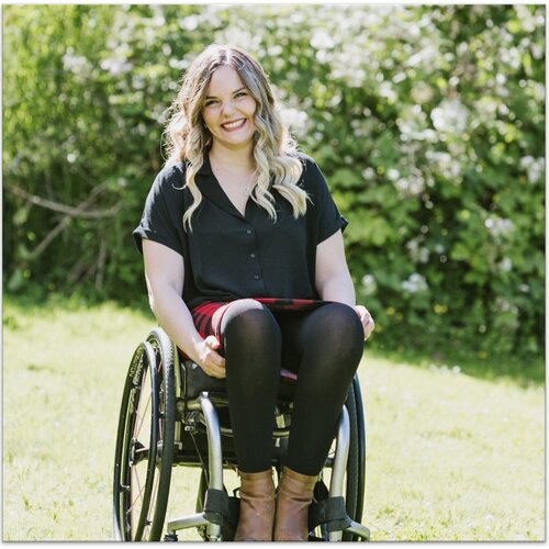 A woman with wavy blonde hair smiles at the camera while sitting in a wheelchair. She is wearing a black top, black leggings, and brown boots. The background features lush greenery, and she is positioned on a grassy area.
