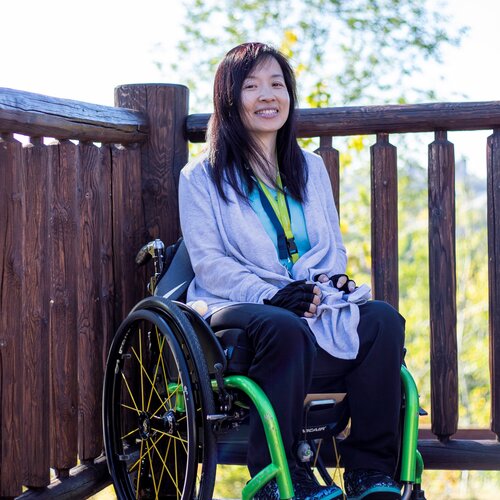 A woman with long dark hair, wearing a gray cardigan and black pants, sits in a bright green wheelchair on a wooden deck with a grassy landscape visible in the background. She is smiling.