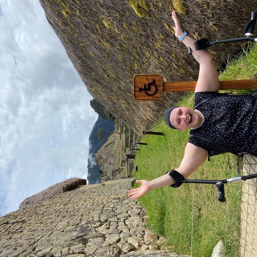 A person sitting on the ground with their arms raised in joy, using crutches. They are on an accessible pathway with a wheelchair access sign beside them. The background shows stone walls and a mountainous landscape. It appears to be a historic or archaeological site.