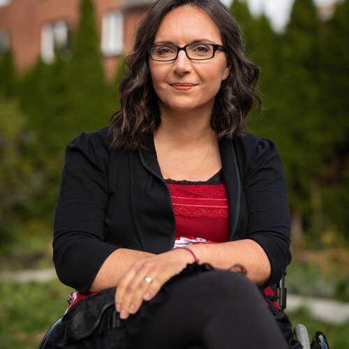 A woman with glasses, wearing a black cardigan over a red top and black pants, is sitting in a wheelchair outdoors. She has dark shoulder-length hair and is looking at the camera with a slight smile. The background shows green shrubbery and a building with windows.