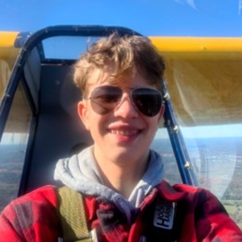 A person wearing sunglasses and a red plaid shirt with a gray hooded sweatshirt underneath smiles while taking a selfie in an airplane cockpit. The background shows a blue sky and landscape seen through the airplane windows.