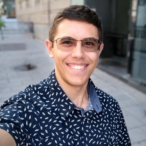 A person wearing sunglasses and a blue patterned shirt is smiling at the camera. The person is standing on a sidewalk with buildings in the background.