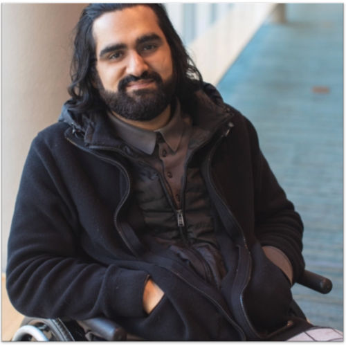 A man with shoulder-length dark hair and a beard is sitting in a wheelchair. He is wearing a black jacket over a dark shirt. He is inside a building with light walls and a blue carpet. He has a calm expression and is looking at the camera.