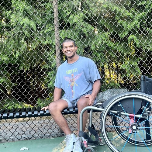 A man in a wheelchair sits on a tennis court in front of a fence with trees in the background. He is wearing a grey Nirvana T-shirt, shorts, and sneakers, and is smiling warmly at the camera.