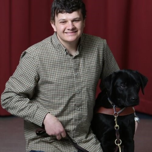 A young man with short brown hair sits in front of a red curtain, smiling and holding the harness of a black Labrador guide dog seated next to him. He wears a green and brown checkered shirt.