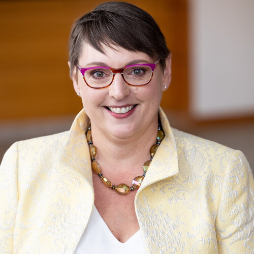 Jane has short brown hair and is wearing purple glasses, a yellow-jeweled necklace, and a yellow collared jacket with white floral patterns.