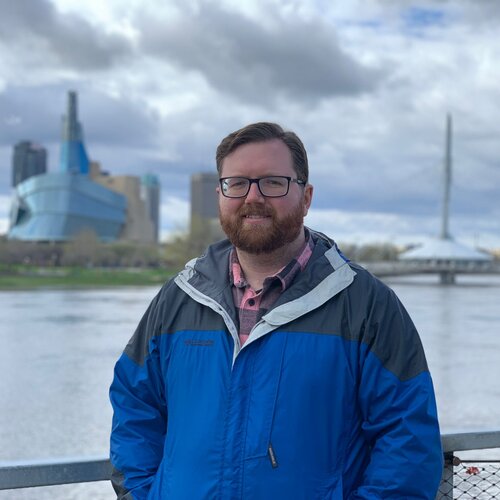 Man in blue rain jacket standing in front of a wide river in an urban setting. 