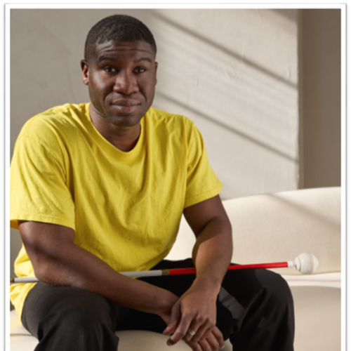 Young man with a serious expression leaning forward with his hands and a white identification cane resting on his knees.