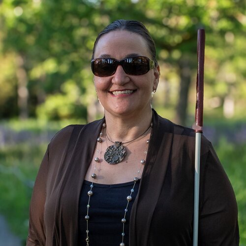 smiling middle aged woman with statement jewlery and outdoors, wearing sunglasses and using a white identification cane