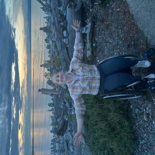 Young woman with arms outspread using a wheelshiar beside a beach at sunset, smiling soflty 