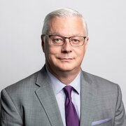 Headshot, middle aged man with white hair, glasses, smiling in grey suit. 